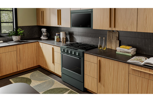 Una escena de cocina moderna que presenta una estufa de gas independiente de color verde oscuro y un microondas empotrado, ubicados entre gabinetes de madera de roble claro con encimeras y protector contra salpicaduras de color gris oscuro.