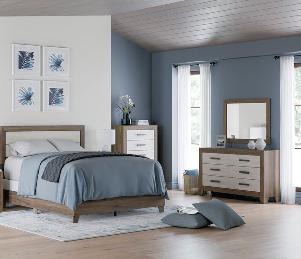 Dormitorio decorado con cama, mesita de noche, cómoda y espejo en un acabado coordinado de roble claro y blanco roto, ubicado frente a paredes azul grisáceo y cortinas blancas. Un ambiente acogedor, luminoso, elegante y de estilo contemporáneo.
