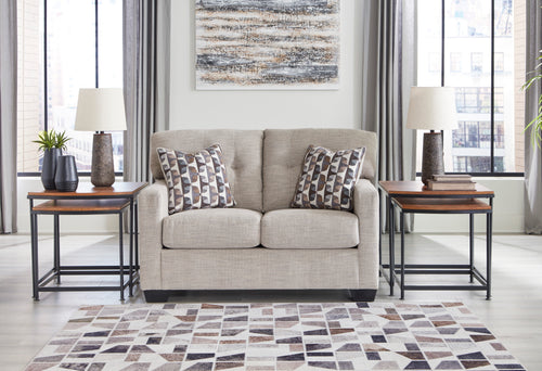 Loveseat de color beige con cojines decorativos y con elementos decorativos en una sala con ventanas en el fondo. 