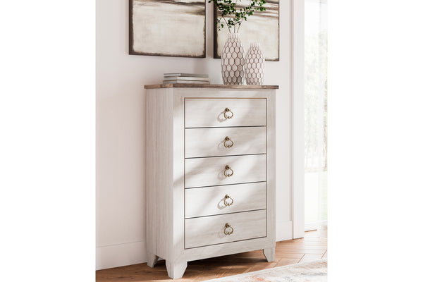 Un chest alto blanco de cinco cajones con superficie de madera marrón clara y tiradores redondos de latón, decorado en una habitación luminosa junto a una ventana.
