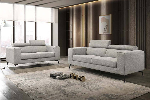 Una sala de estar de estilo moderno que cuenta con un sofá de dos plazas y otro de tres plazas a juego —ambos de color gris claro y con patas finas de metal negro—, situados sobre una alfombra beige texturizada frente a una pared revestida de paneles de madera oscura.