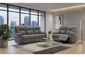 Un moderno salón con un sofá y loveseat reclinable tapizado en gris a juego, dispuestos sobre una alfombra estampada con una mesa de centro de cristal entre ellos. La habitación cuenta con una pared con textura de hormigón, un gran ventanal con vistas al horizonte de la ciudad y plantas de interior.