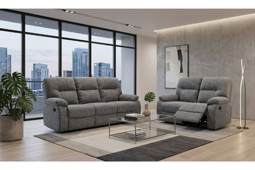 Un moderno salón con un sofá y loveseat reclinable tapizado en gris a juego, dispuestos sobre una alfombra estampada con una mesa de centro de cristal entre ellos. La habitación cuenta con una pared con textura de hormigón, un gran ventanal con vistas al horizonte de la ciudad y plantas de interior.