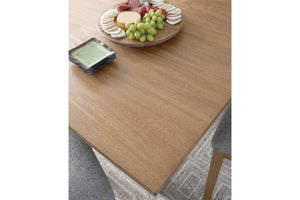 Una mesa de comedor de madera en tono café mediano cálido, mostrada en un ambiente interior decorado con una tabla de charcutería, uvas, galletas saladas y un plato pequeño. En la parte inferior derecha se observa parte de una silla tapizada en color gris.