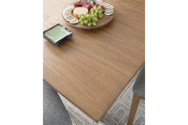 Una mesa de comedor de madera en tono café mediano cálido, mostrada en un ambiente interior decorado con una tabla de charcutería, uvas, galletas saladas y un plato pequeño. En la parte inferior derecha se observa parte de una silla tapizada en color gris.