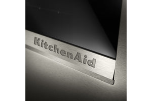Primer plano de un embellecedor de acero inoxidable cepillado para un electrodoméstico KitchenAid, con el logotipo de KitchenAid grabado en relieve sobre un panel de vidrio oscuro.