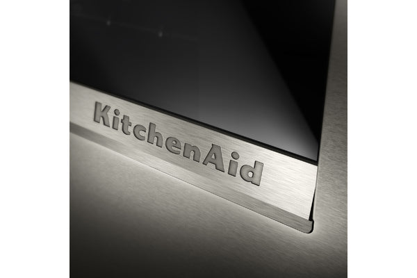 Primer plano de un embellecedor de acero inoxidable cepillado para un electrodoméstico KitchenAid, con el logotipo de KitchenAid grabado en relieve sobre un panel de vidrio oscuro.