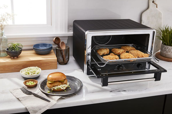 Un horno de convección/tostador de sobremesa en una cocina luminosa, con la puerta abierta y una cesta de alambre con trozos de pollo frito dorado en su interior. Una hamburguesa y unos pequeños acompañamientos reposan sobre la encimera, cerca de allí.