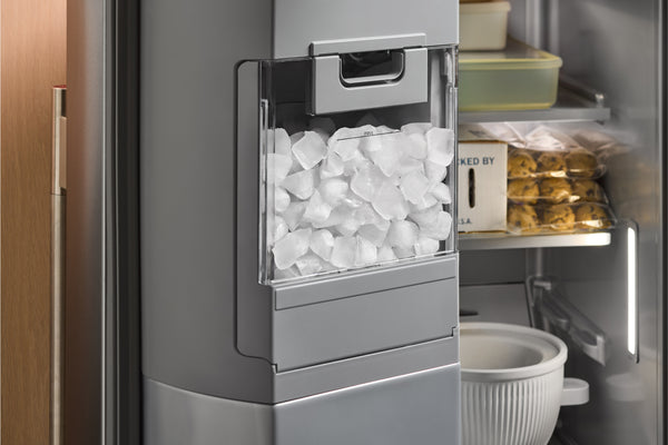 Primer plano de la puerta de un refrigerador de acero inoxidable con el cajón dispensador de hielo abierto, lleno de cubitos de hielo, y los estantes interiores visibles detrás.