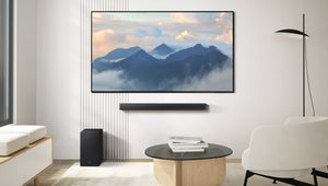Sala de estar moderna con televisor de pantalla plana montado en la pared sobre una barra de sonido y un subwoofer. Incluye mesa de centro redonda, sillón blanco y lámpara de pie, con una imagen de montañas en la pantalla del televisor.