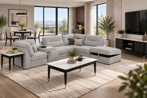 Un moderno sofá modular de color gris claro con chaise longue en forma de L, situado en una sala de estar contemporánea y luminosa con mesa de centro blanca, alfombra texturizada y grandes ventanales.