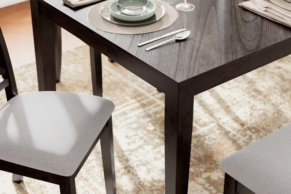 Mesa de comedor de madera oscura con sillas tapizadas en gris en un ambiente moderno, decorada con vajilla y una alfombra con diseño.