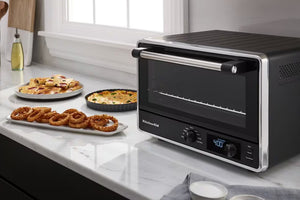 Un horno de convección de sobremesa KitchenAid, de color negro y acero inoxidable, descansa sobre una encimera de mármol blanco junto a platos de comida, entre los que se incluyen aros de cebolla, pizza y un pastel horneado.