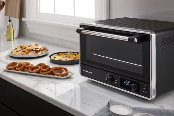 Un horno de convección de sobremesa KitchenAid, de color negro y acero inoxidable, descansa sobre una encimera de mármol blanco junto a platos de comida, entre los que se incluyen aros de cebolla, pizza y un pastel horneado.