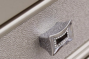 Detalle en primer plano de una gaveta de mueble con acabado texturizado en tono beige claro y tirador metálico plateado con incrustaciones brillantes. La superficie presenta relieve fino tipo grano, destacando un diseño elegante y decorativo.