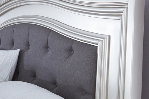 Vista cercana de una cama tapizada y plateada con acolchado blanco y con cabecera gris oscura sobre un fondo blanco. 