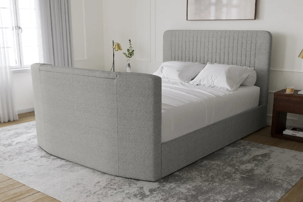 Una cama tapizada de estilo moderno con cabecero curvo y piecero a juego, presentada en un dormitorio luminoso con ropa de cama blanca, una alfombra gris y una mesita de noche de madera.