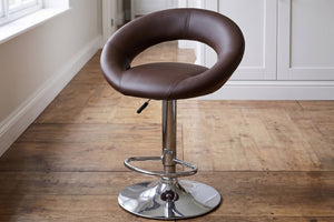 Taburete de bar ajustable en color marrón con asiento acolchado y respaldo curvo envolvente. Cuenta con reposapiés y base cromada redonda. Se muestra en un ambiente de cocina moderna con piso de madera.