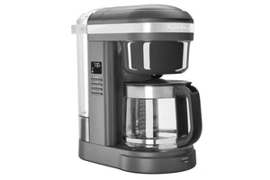 Una cafetera KitchenAid en color gris oscuro y acero inoxidable, con pantalla digital, jarra de vidrio con asa negra y depósito de agua transparente, mostrada sobre un fondo blanco liso.