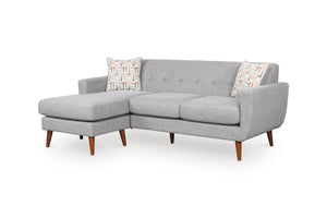 Sofa chaise de color gris claro con patas acabadas en madera y con dos cojines decorativos sobre un fondo blanco. 