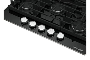 Primer plano de un tope de gas KitchenAid negra que muestra cinco perillas de control plateadas y parte de la rejilla de hierro fundido negra sobre los quemadores sobre un fondo blanco.
