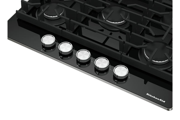 Primer plano de un tope de gas KitchenAid negra que muestra cinco perillas de control plateadas y parte de la rejilla de hierro fundido negra sobre los quemadores sobre un fondo blanco.