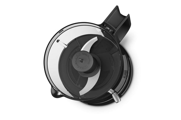 Vista superior de la tapa de un electrodoméstico de cocina negro, probablemente la tapa del recipiente de una procesadora de alimentos o una batidora, con un borde de plástico transparente y una carcasa central negra para la cuchilla con un accesorio plateado para la misma, sobre un fondo blanco liso.
