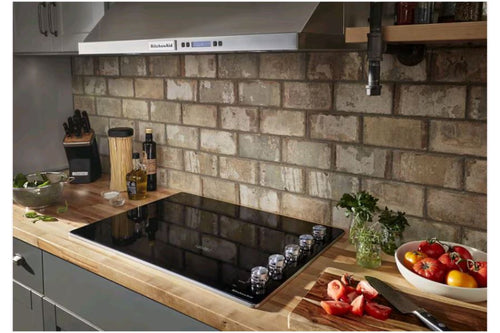 Cocina moderna con estufa empotrable KitchenAid de superficie vitrocerámica negra sobre tope de madera. Incluye controles frontales, campana extractora de acero inoxidable y área de preparación con vegetales frescos.