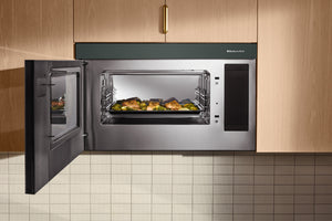 Un horno microondas empotrado de acero inoxidable y color verde, con la puerta abierta, que deja ver una bandeja de comida asada en su interior, situado entre armarios de cocina de madera clara sobre una encimera de azulejos.