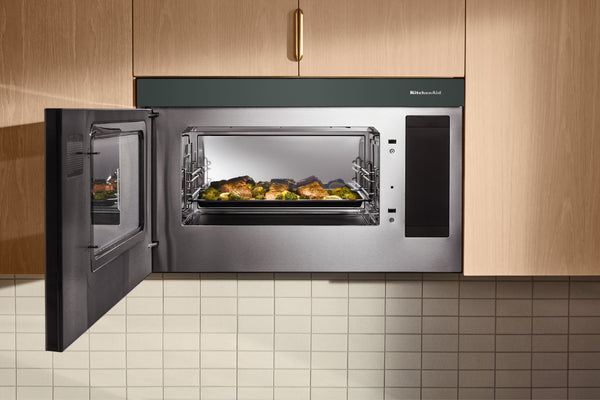 Un horno microondas empotrado de acero inoxidable y color verde, con la puerta abierta, que deja ver una bandeja de comida asada en su interior, situado entre armarios de cocina de madera clara sobre una encimera de azulejos.