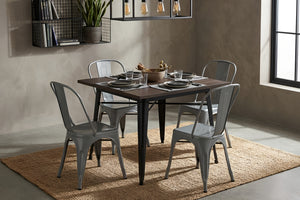 Mesa de comedor cuadrada con superficie de madera oscura y base metálica negra, acompañada por cuatro sillas grises de estilo industrial. El conjunto está dispuesto sobre una alfombra tejida en un espacio moderno con iluminación colgante.