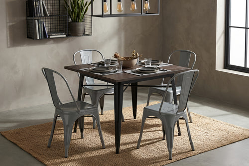 Mesa de comedor cuadrada con superficie de madera oscura y base metálica negra, acompañada por cuatro sillas grises de estilo industrial. El conjunto está dispuesto sobre una alfombra tejida en un espacio moderno con iluminación colgante.