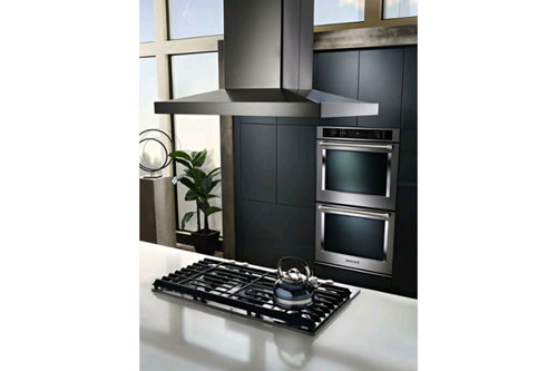 Cocina moderna con campana extractora KitchenAid de acero inoxidable instalada sobre una estufa de gas empotrable. El espacio incluye hornos de pared integrados y acabados contemporáneos en tonos oscuros y metálicos.