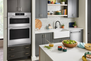 Un interior de cocina moderno que cuenta con un horno doble empotrado en la pared, con gabinetes de color gris oscuro, acero inoxidable y frentes negros, junto a un fregadero estilo granja, un protector contra salpicaduras de azulejos blancos tipo metro, estantes abiertos con utensilios de cocina y una isla blanca en primer plano con ensaladas y alimentos.