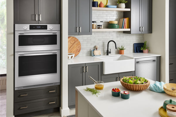 Un interior de cocina moderno que cuenta con un horno doble empotrado en la pared, con gabinetes de color gris oscuro, acero inoxidable y frentes negros, junto a un fregadero estilo granja, un protector contra salpicaduras de azulejos blancos tipo metro, estantes abiertos con utensilios de cocina y una isla blanca en primer plano con ensaladas y alimentos.