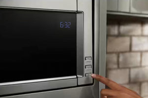 Primer plano de un horno microondas de acero inoxidable con puerta negra y pantalla digital azul que muestra la hora, donde se ve un dedo pulsando los botones de control del lado derecho.