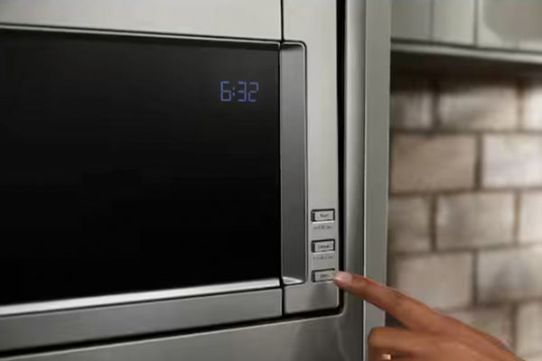 Primer plano de un horno microondas de acero inoxidable con puerta negra y pantalla digital azul que muestra la hora, donde se ve un dedo pulsando los botones de control del lado derecho.