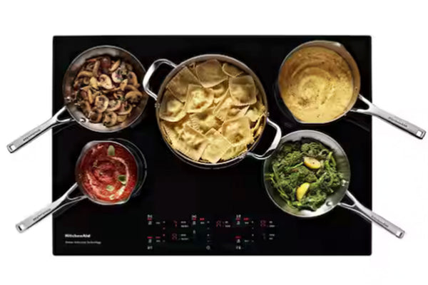 Vista superior de una placa de inducción negra con cinco sartenes de acero inoxidable cocinando diferentes alimentos, como pasta, champiñones, sopa, salsa de tomate y verduras, sobre un fondo blanco.