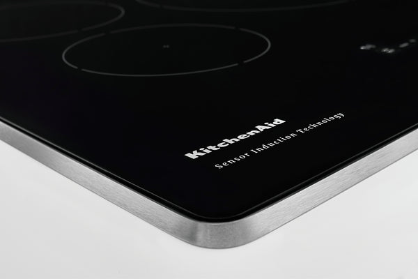 Primer plano de un tope de inducción negro con esquinas redondeadas, un borde de metal plateado cepillado y el logotipo de KitchenAid impreso en la superficie.