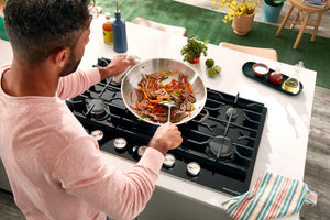 Un hombre con camisa rosa cocina en una sartén de acero inoxidable sobre una estufa de gas negra instalada en una isla de cocina blanca. La escena está bien iluminada y, al fondo, se aprecia la estética de una cocina moderna al aire libre.