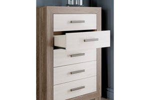 Chest de 5 cajones con estructura de madera de roble claro y frentes de cajón en beige claro, con tiradores metálicos plateados. Un cajón se muestra ligeramente abierto, sobre un fondo de pared gris oscuro.