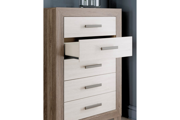 Chest de 5 cajones con estructura de madera de roble claro y frentes de cajón en beige claro, con tiradores metálicos plateados. Un cajón se muestra ligeramente abierto, sobre un fondo de pared gris oscuro.