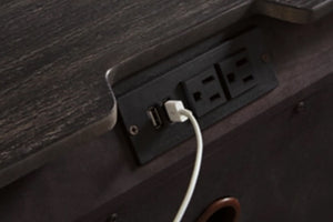 Primer plano de una superficie de madera oscura con una regleta negra integrada que incluye un puerto USB y dos enchufes de pared, con un cable de carga blanco conectado.