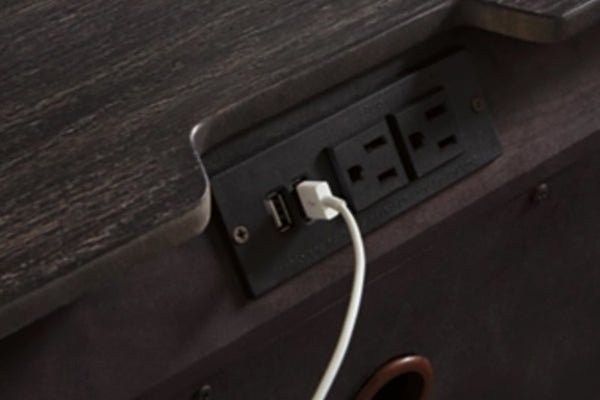 Primer plano de una superficie de madera oscura con una regleta negra integrada que incluye un puerto USB y dos enchufes de pared, con un cable de carga blanco conectado.