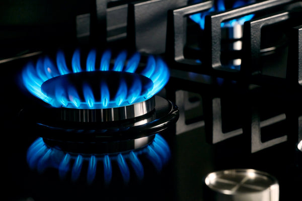 Primer plano de un quemador de estufa de gas encendido con llamas de color azul brillante, sobre una encimera de color negro oscuro con rejillas metálicas.