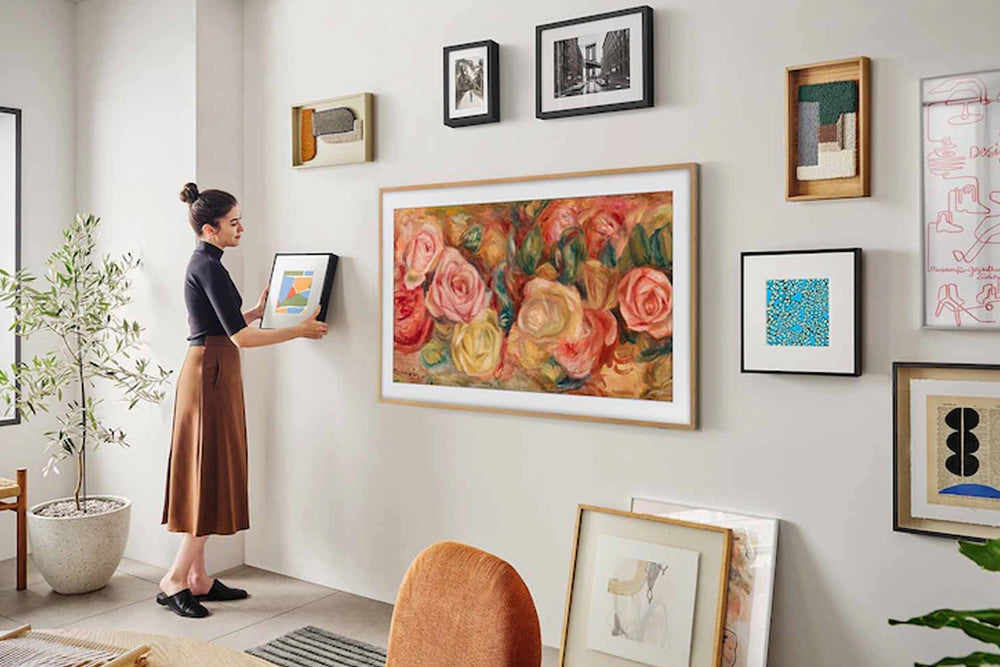 Mujer organizando obras de arte enmarcadas con un televisor en el medio de una pared en un entorno doméstico.