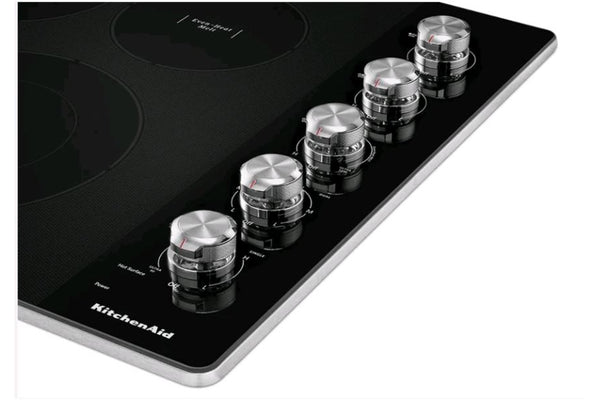 Primer plano de los controles giratorios de una estufa empotrable KitchenAid con superficie vitrocerámica negra. Perillas metálicas de precisión para ajustar la temperatura de cinco zonas de cocción eléctricas.