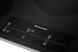 Primer plano de un tope de inducción KitchenAid negro con un panel de control brillante e iconos blancos impresos sobre una superficie oscura texturizada.