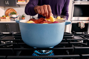 Una persona con una camisa morada añade una patata a una olla de hierro fundido esmaltada de color azul claro que se encuentra sobre una estufa de gas en una cocina.