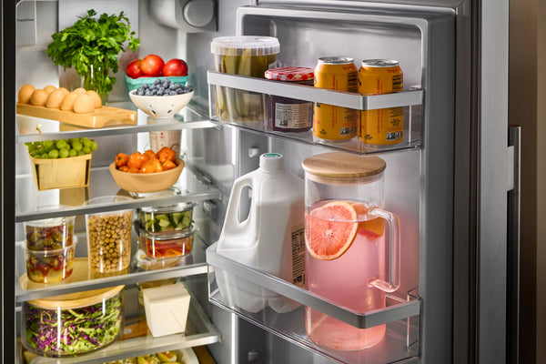 Interior abierto de acero inoxidable con estantes llenos de verduras frescas, frutas, recipientes y una jarra de leche en la puerta, además de una jarra de vidrio llena de bebida rosa y rodajas de pomelo.